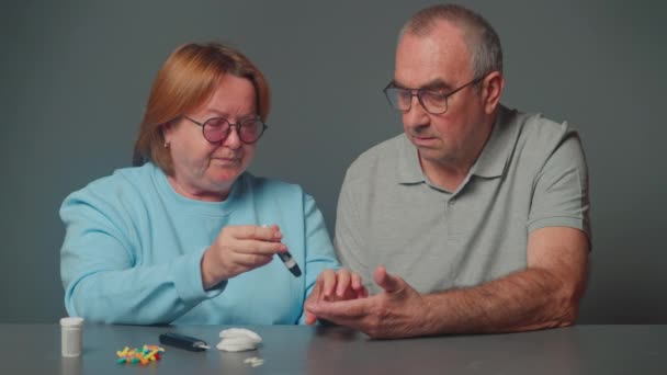 テーブルに座っている老夫婦 妻は彼女の糖尿病の夫が交換可能な針の携帯用接着剤を使用して迅速な血糖検査を行うのを助けます — ストック動画
