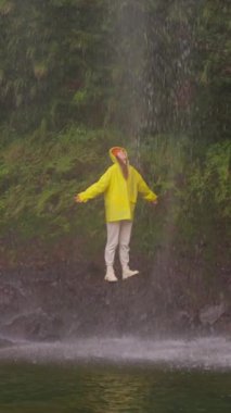  Dikey video. Şelale altında kollarını açmış, doğa macerasında yürüyüş yapan bir kadın açık hava tatilinde keşfe çıkıyor. Seyahat, özgürlük ya da yolculuk.