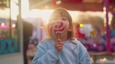 Eğlence parkında gülümseyen küçük kız. Elinde büyük bir çubuk şeker olan çocuk gülümser ve çerçevede duygularını gösterir, yanan atlıkarıncaların arka planında durur..