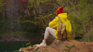 Sarı ceketli ve sırt çantalı bir kadın gezgin ulaşılması zor güzel bir şelaleye ulaştı ve oturur, hayran kalır ve güzelliğinin tadını çıkarır. Yüksek kalite 4k görüntü