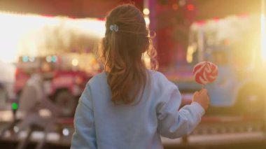 Küçük sevimli bir kız elinde büyük çizgili bir lolipop tutuyor ve gün batımında bir eğlence parkında ışıldayan bir cazibeye bakıyor..