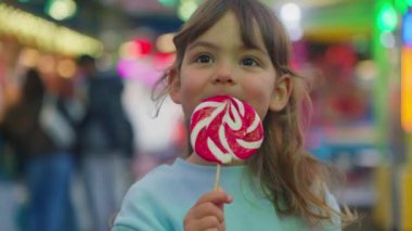 Küçük, güzel bir kız elinde büyük çizgili bir lolipop tutuyor ve onu eğlence parkında parlayan arabaların arka planında yiyor. Yüksek kalite 4k görüntü