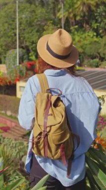 Dikey video. Gezgin, Madeira ve Funchal şehrinin çeşitli bitki örtüleriyle birlikte tarihi bir bahçeyi ziyaret ediyor. Botanik bahçesinde renkli çiçekler arasında yürüyen şapkalı ve sırt çantalı bir kadın..