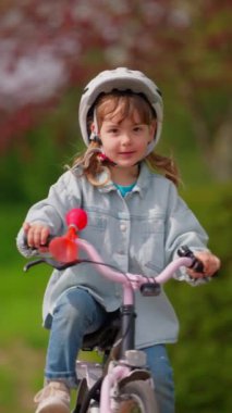 Dikey video. Beyaz kasklı şirin küçük bir kız bisiklet sürüyor gülümsüyor ve arka plandaki stadyumda mutlu hissediyor. Çocuklar için spor etkinliği.