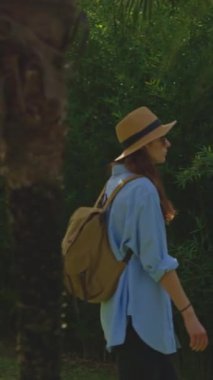 Dikey video. Kamera, şapkalı ve sırt çantalı gezgin kadını takip ediyor. Ormandaki tropikal bahçede ağır çekim kadın yürüyüşü..