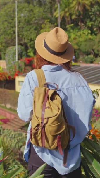 バーティカルビデオ マデイラ島とファチュアルシティの多様な植生を持つランドマークガーデンを訪れる旅行者 植物園のカラフルな花の間を歩く帽子とバックパックを持つ女性 — ストック動画