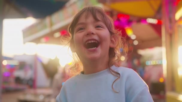 一个漂亮的小女孩手里拿着一个大的条纹棒棒糖 在游乐园里光彩夺目的游乐设施前吃着它 高质量的4K镜头 — 图库视频影像