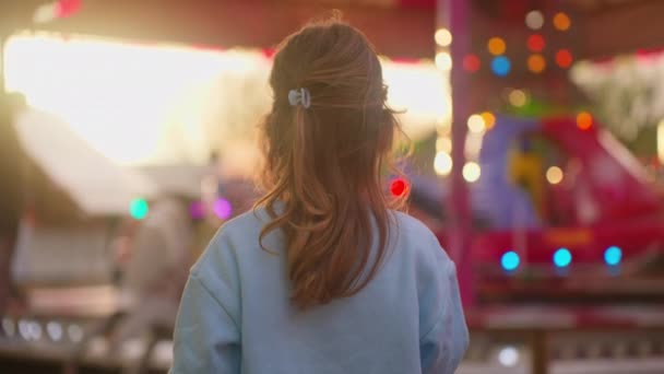 一个可爱的小女孩手里拿着一个大的条纹棒棒糖 在夕阳西下的游乐园里观看着一个发光的景点 — 图库视频影像