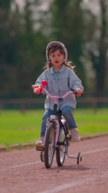 Dikey video. Beyaz kasklı şirin küçük bir kız bisiklet sürüyor gülümsüyor ve arka plandaki stadyumda mutlu hissediyor. Çocuklar için spor etkinliği.