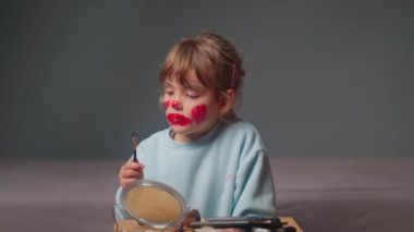Aynanın karşısında oturan tatlı küçük bir kız makyaj yapmak için kozmetik ürünlerine fırça batırmaya çalışıyor..