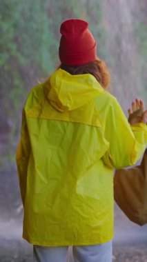 Dikey video. Turistik kıyafetli, sarı ceketli ve sırt çantalı bir kadın şelalenin inanılmaz manzarasına bakarak destansı manzaranın üzerinde yürüyor..