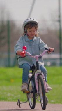 Dikey video. Beyaz kasklı şirin küçük bir kız bisiklet sürüyor gülümsüyor ve arka plandaki stadyumda mutlu hissediyor. Çocuklar için spor etkinliği.