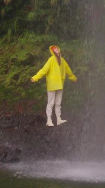 Dikey video. Şelale altında kollarını açmış, doğa macerasında yürüyüş yapan bir kadın açık hava tatilinde keşfe çıkıyor. Seyahat, özgürlük ya da yolculuk. 