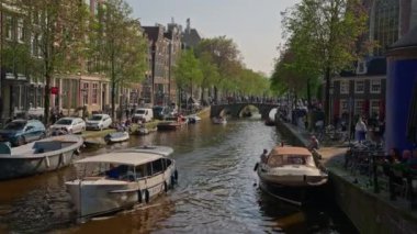 Amsterdam, Hollanda, 09 Haziran 2024: Kanal boyunca geçen turist teknesi, köprüsü ve eski evleri ile Amsterdam manzaralı şehir manzaralı bir kanalın kuruluşu. İnsanlar bisiklet ve elektrikli tekne kullanıyor..