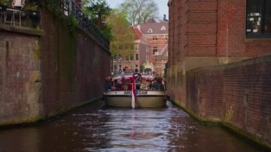 Amsterdam, Hollanda, 09 Haziran 2024: Kanal boyunca geçen turist teknesi, köprüsü ve eski evleri ile Amsterdam manzaralı şehir manzaralı bir kanalın kuruluşu. İnsanlar bisiklet ve elektrikli tekne kullanıyor..
