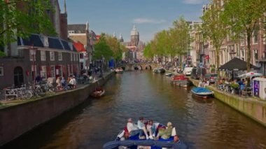 Amsterdam, Hollanda, 09 Haziran 2024: Kanal boyunca geçen turist teknesi, köprüsü ve eski evleri ile Amsterdam manzaralı şehir manzaralı bir kanalın kuruluşu. İnsanlar bisiklet ve elektrikli tekne kullanıyor..