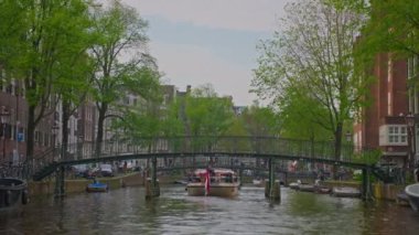 Amsterdam, Hollanda, 09 Haziran 2024: Kanal boyunca geçen turist teknesi, köprüsü ve eski evleri ile Amsterdam manzaralı şehir manzaralı bir kanalın kuruluşu. İnsanlar bisiklet ve elektrikli tekne kullanıyor..