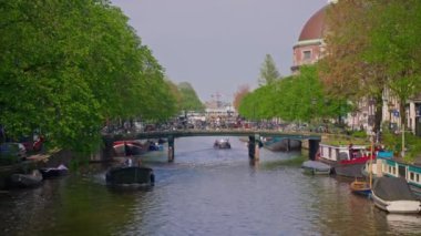 Amsterdam, Hollanda, 09 Haziran 2024: Kanal boyunca geçen turist teknesi, köprüsü ve eski evleri ile Amsterdam manzaralı şehir manzaralı bir kanalın kuruluşu. İnsanlar bisiklet ve elektrikli tekne kullanıyor..