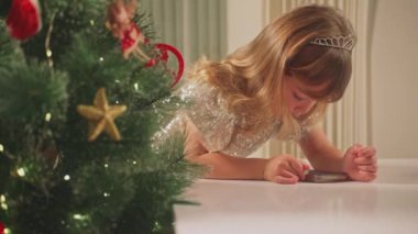 Gülümseyen küçük bir kız, ışıl ışıl bir Noel ağacının zemininde telefonla oynuyor..