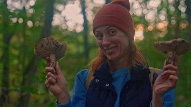 Turuncu şapkalı mutlu bir kadının portresi ve ormanda elinde büyük mantarlarla onları kameraya gösteriyor..