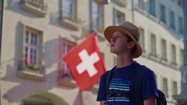 Eski Bern şehrinin merkezinde sırt çantalı ve şapkalı genç turist öğrenci mimariyi izliyor, İsviçre bayrağının arka planında binalar var. İsviçre.
