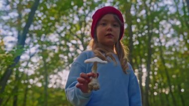 Büyük bir mantar tutan küçük güzel bir kızın portresi ve sonbaharda bir ormanın arka planında kameraya mantarı gösteriyor..