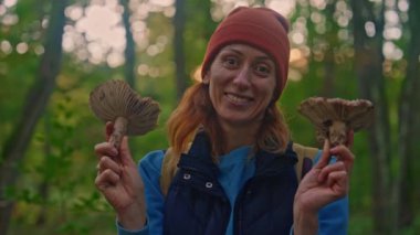 Turuncu şapkalı mutlu bir kadının portresi ve ormanda elinde büyük mantarlarla onları kameraya gösteriyor..