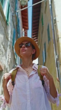 Dikey video. Sırt çantalı genç ve çekici bir turist seyahat ederken eski şehrin sokaklarında geziniyor, mimariye hayran, etrafa bakıyor. İtalya.