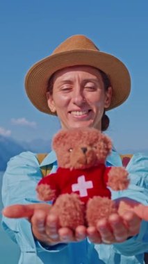 dikey video. Şapkalı bir kadın, Brienz şehrindeki Interlaken gölünün arka planında İsviçre bayrağı şeklinde küçük yumuşak bir oyuncak ayı tutuyor..