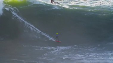 Sörfçü büyük dalgalarda sörf yapıyor. Yanında da yakınlarda bir jet ski var. Aşırı büyük dalga sörfü. Nazare, Portekiz.