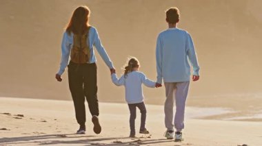 Çocuklarıyla birlikte bir anne, genç bir oğlan ve küçük bir kız gün batımında okyanus kıyısında yürür..