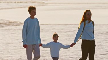 Çocuklarıyla birlikte bir anne, genç bir oğlan ve küçük bir kız gün batımında okyanus kıyısında yürür. Aile el ele tutuşuyor..