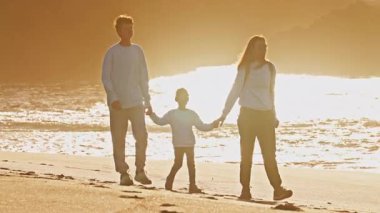 Çocuklarıyla birlikte bir anne, genç bir oğlan ve küçük bir kız gün batımında okyanus kıyısında yürür. Aile el ele tutuşuyor..