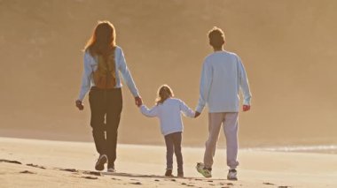 Çocuklarıyla birlikte bir anne, genç bir oğlan ve küçük bir kız gün batımında okyanus kıyısında yürür..