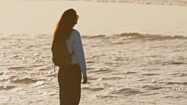 Sırt çantalı kız okyanus kıyısında duruyor ve altın saatinde günbatımının tadını çıkarıyor..