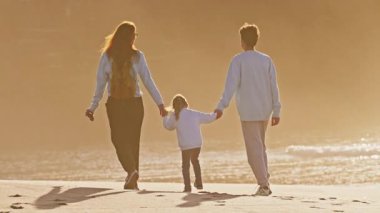 Çocuklarıyla birlikte bir anne, genç bir oğlan ve küçük bir kız gün batımında okyanus kıyısında yürür. Aile el ele tutuşuyor..