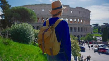 Şapkalı ve sırt çantalı bir kız, güneşli bir günde Roma 'daki Colosseum' a bakıyor..