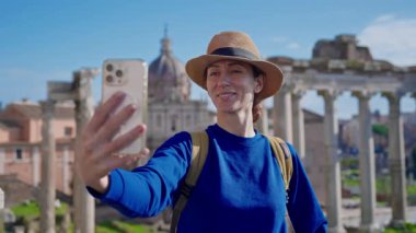 Şapkalı ve sırt çantalı mutlu bir turist kadın, Roma Forumu 'ndaki Antik Roma Sütunları' nın manzara arkaplanının önünde telefonuyla selfie çekiyor. İtalya.