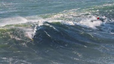 Sörfçü büyük dalgalarda sörf yapıyor. Yanında da yakınlarda bir jet ski var. Aşırı büyük dalga sörfü. Nazare, Portekiz. 