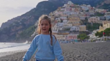 Positano sahilinde durup kollarını sallayarak gülen tatlı bir kız. Sahilden şehrin ana kısmına, dağdaki renkli evlere bakın. Positano, Amalfi Sahili, İtalya.