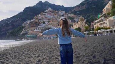 Positano sahilinde durup kollarını sallayarak gülen tatlı bir kız. Sahilden şehrin ana kısmına, dağdaki renkli evlere bakın. Positano, Amalfi Sahili, İtalya.