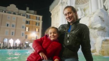 Küçük kızıyla birlikte bir turist kız, Roma 'daki dünyaca ünlü Trevi Çeşmesi' nin arka planında fotograf çekiyor. Çeşmenin aydınlatıldığı akşam. İtalya.