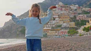 Positano sahilinde dans eden küçük tatlı kız gülüyor. Sahilden şehrin ana kısmına, dağdaki renkli evlere bakın. Positano, Amalfi Sahili, İtalya.
