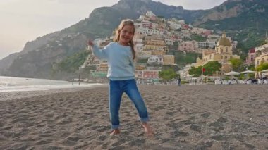 Positano sahilinde dans eden küçük tatlı kız gülüyor. Sahilden şehrin ana kısmına, dağdaki renkli evlere bakın. Positano, Amalfi Sahili, İtalya.