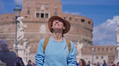 Şapkalı ve sırt çantalı bir turist Tiber nehrinin üzerindeki Ponte Sant Angelo köprüsündeki Aziz Melek Kalesi 'nin arka planında Roma, İtalya' nın manzarasına bakıyor..