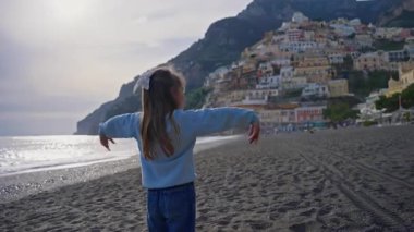 Positano sahilinde durup kollarını sallayarak gülen tatlı bir kız. Sahilden şehrin ana kısmına, dağdaki renkli evlere bakın. Positano, Amalfi Sahili, İtalya.