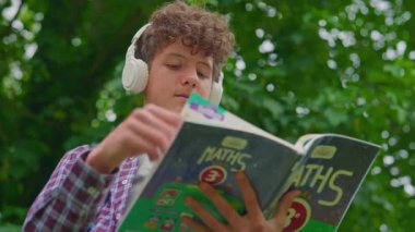 Kıvırcık çocuk kulaklıkla müzik dinliyor ve parkta kitap okuyor..