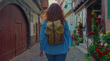 Sırt çantalı bir kadın Eguisheim, Fransa 'nın çiçekleriyle dolu renkli evlerle dolu güzel bir sokakta yürüyor.. 