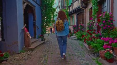 Sırt çantalı bir kadın Eguisheim, Fransa 'nın çiçekleriyle dolu renkli evlerle dolu güzel bir sokakta yürüyor.. 