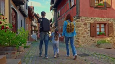 Sırt çantalı bir kadın ve çocuklar Eguisheim, Fransa 'nın çiçekleriyle dolu renkli evlerle dolu güzel bir sokakta yürüyor..